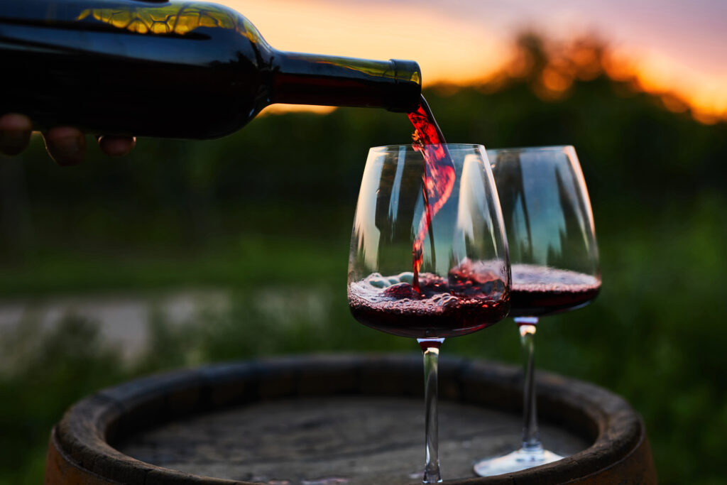 Pouring red wine into glasses on a barrel at dusk.